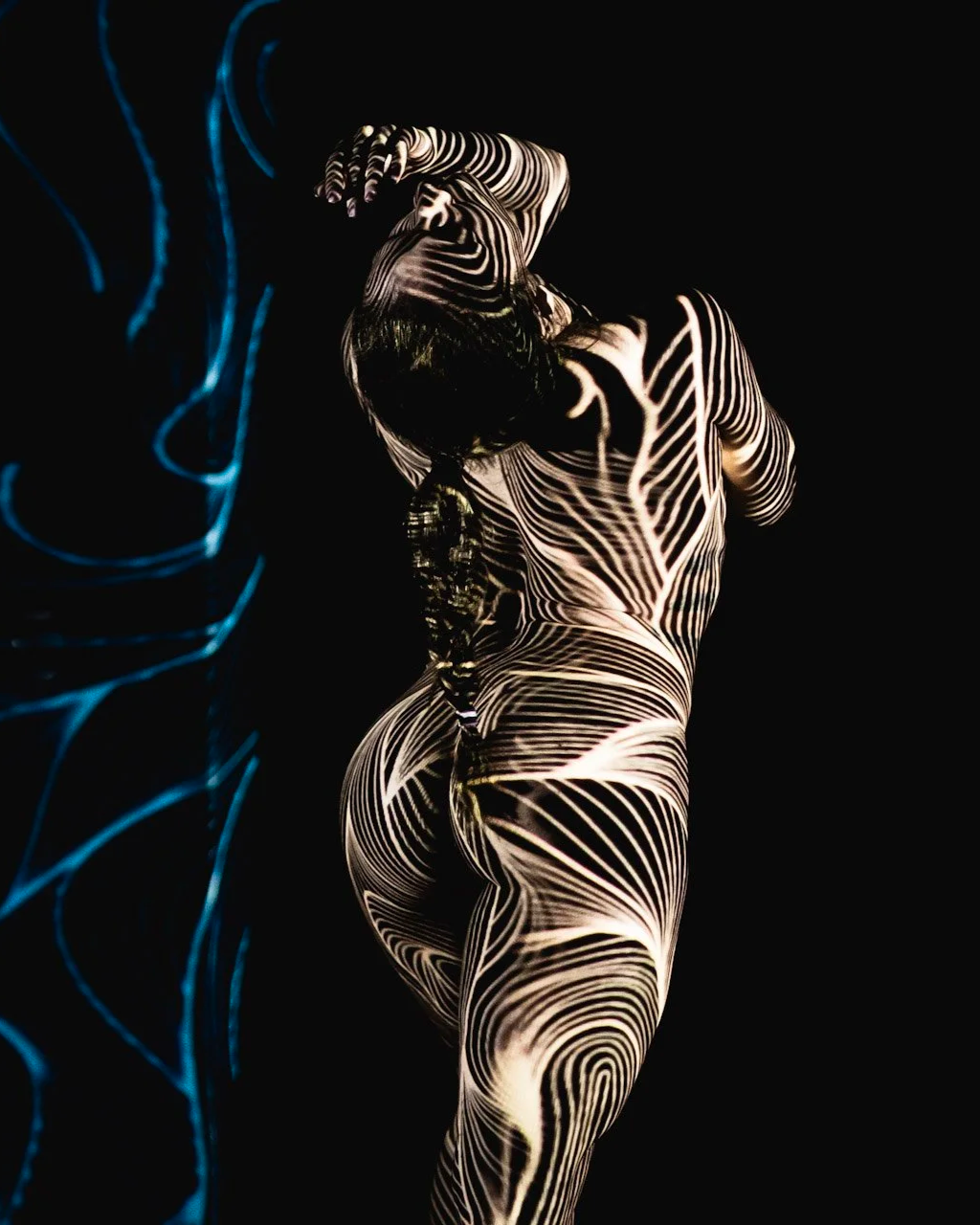 A woman with wet hair posing against a black background, with black and white laser or light patterns projected on her body.
