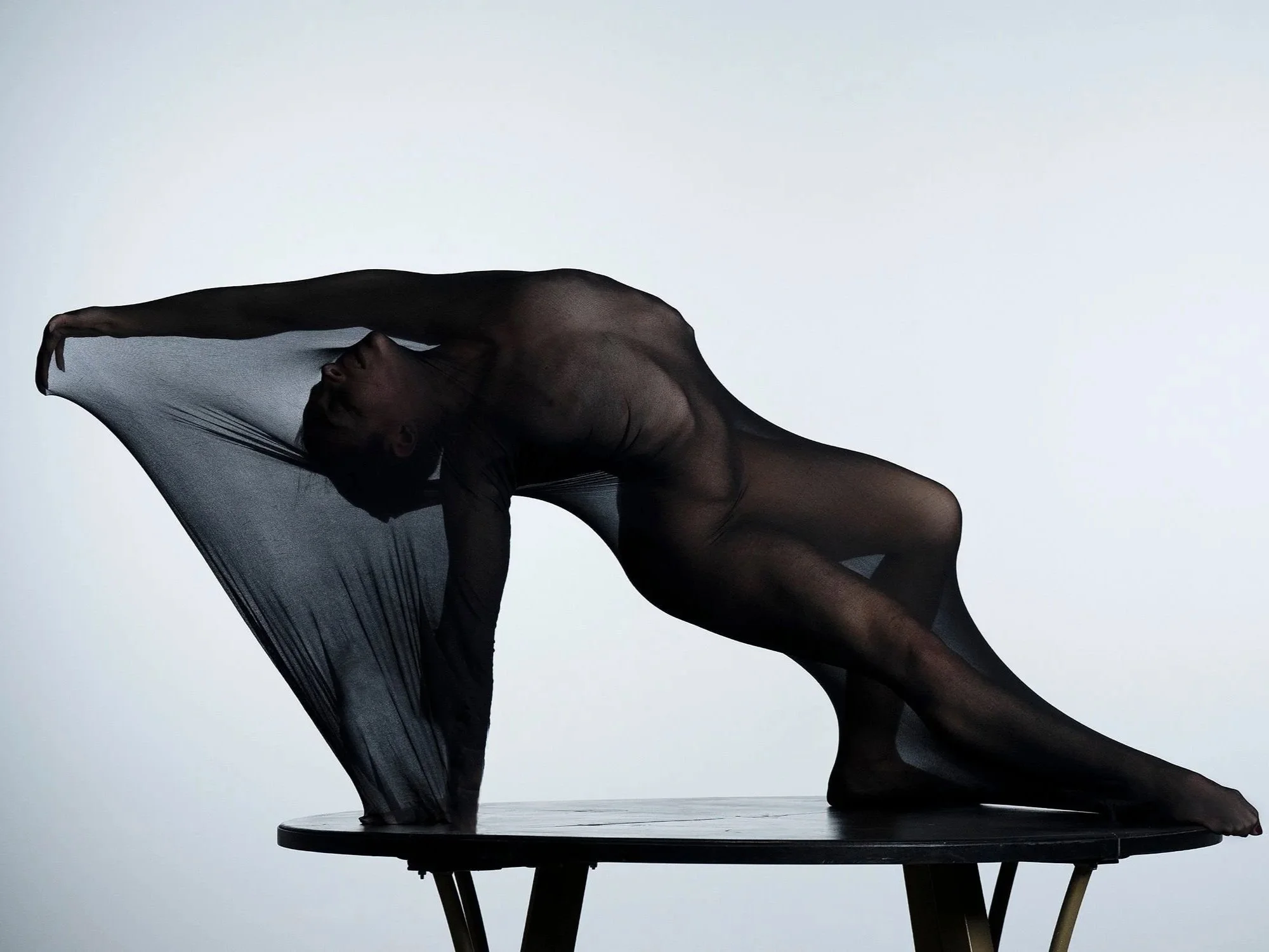 Contortionist draped over a table in a backbend pose with sheer fabric covering the body