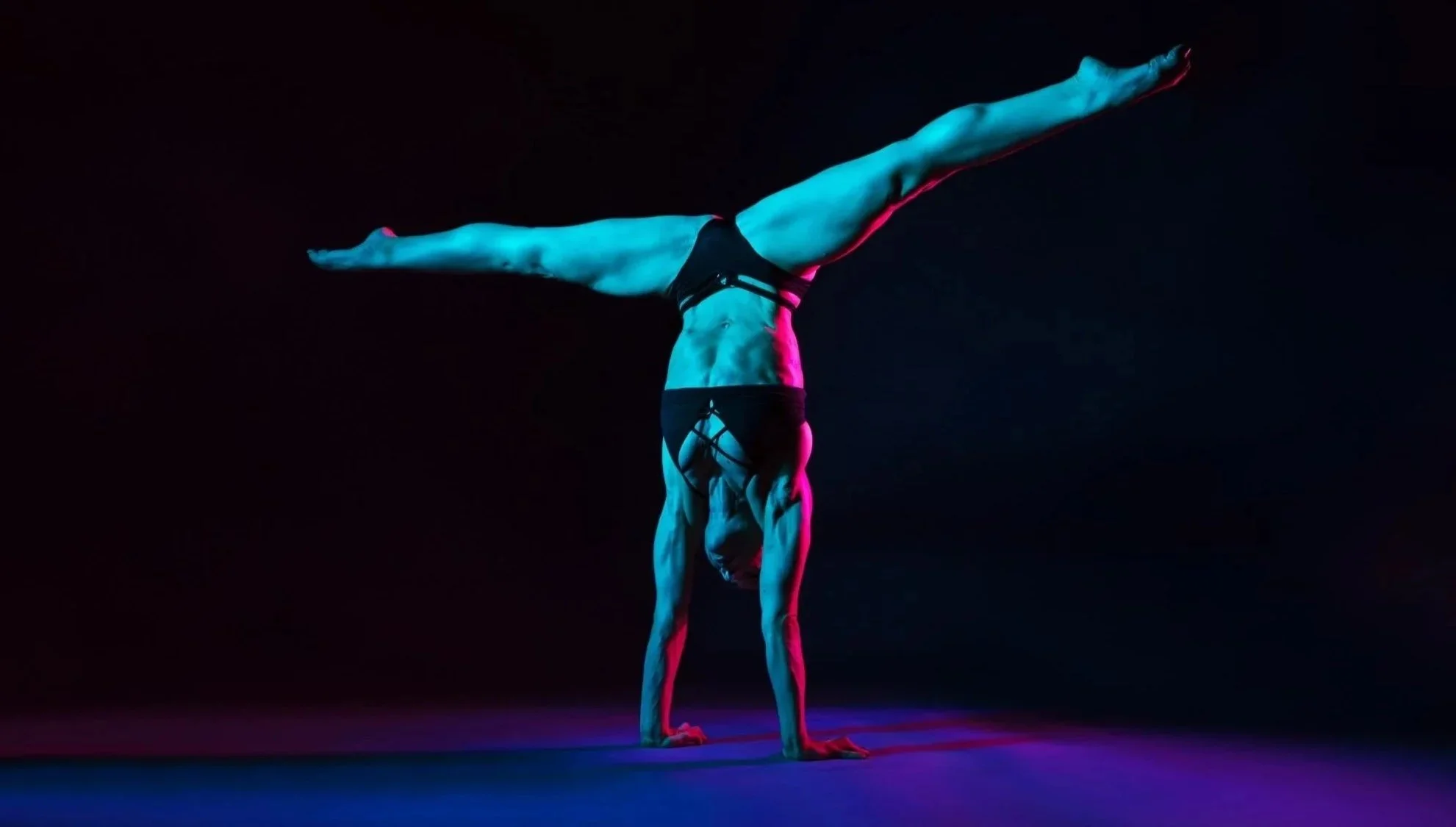 Figure in a straddle handstand under pink and blue studio lighting