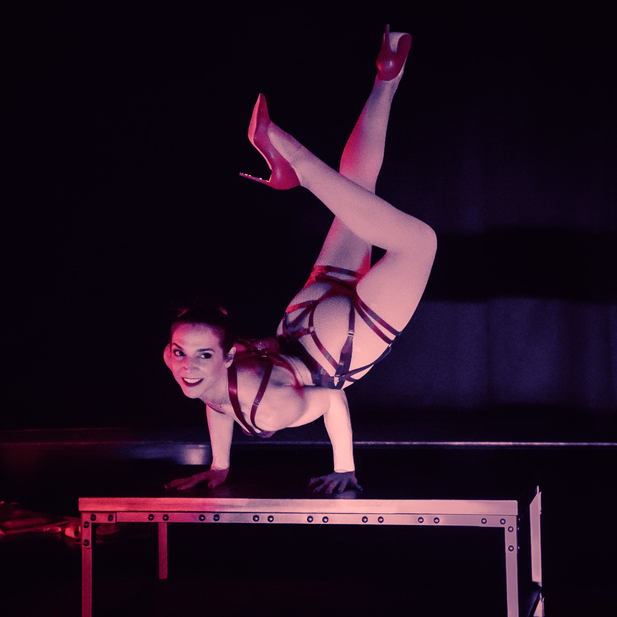 Performer hand balancing on a table with legs extended upward under red and purple lights.