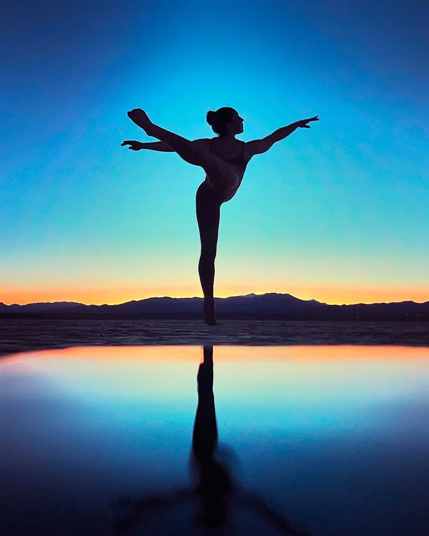 Silhouette of a dancer  balancing on one leg at sunset with arms extended, reflected on water.