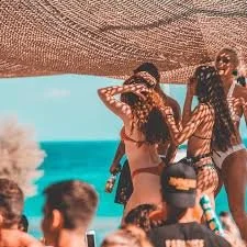 A group of people at a beach or poolside, with some wearing swimsuits and hats, under a shaded area, with the ocean in the background.