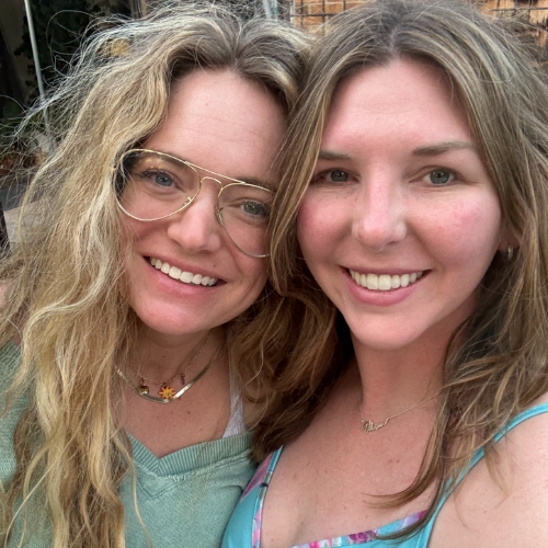 Two smiling women taking a selfie outdoors, one with glasses and the other with long, wavy hair.