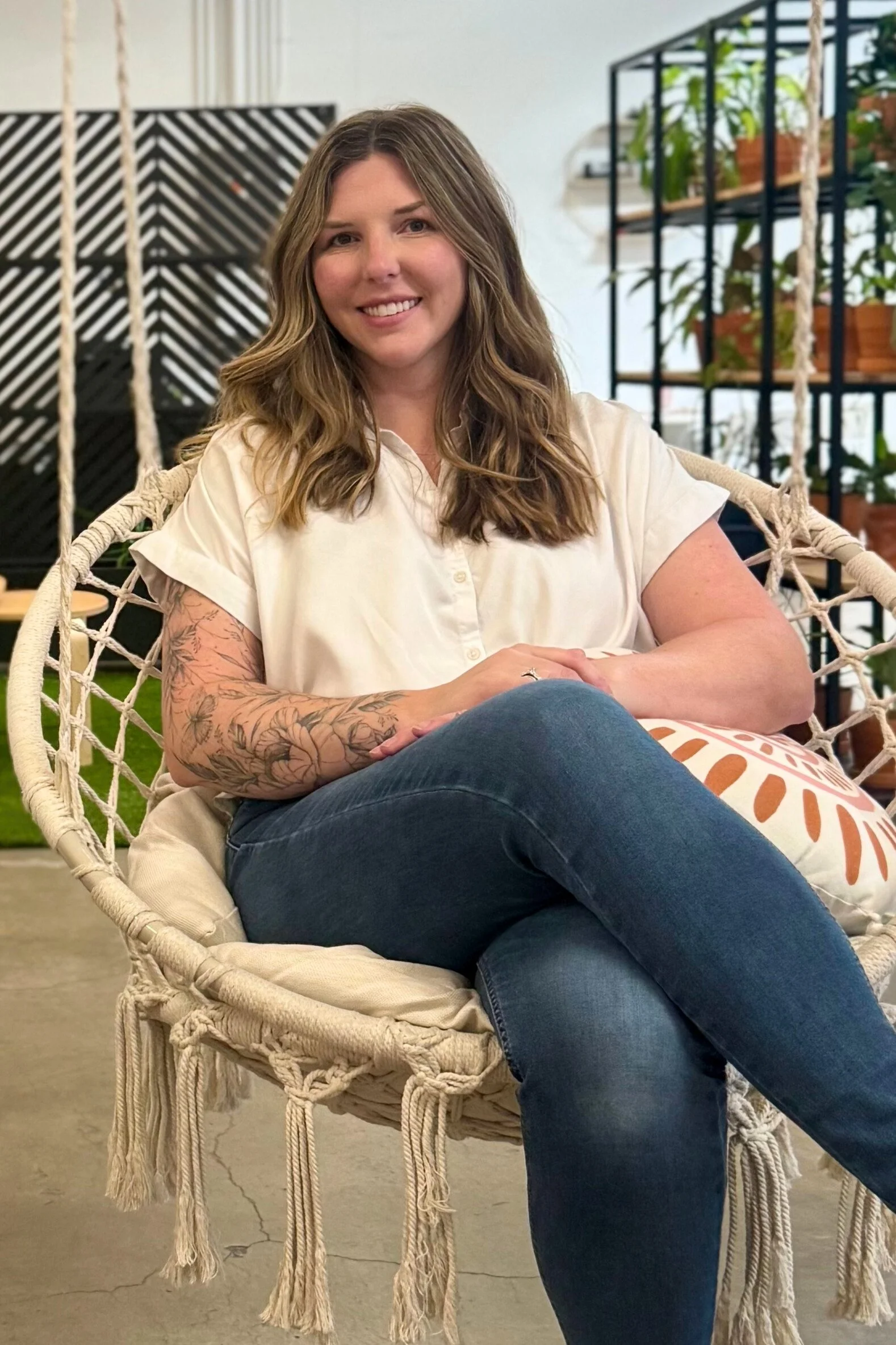 A woman with long, wavy hair, wearing a cream-colored blouse and jeans, sitting on a woven hanging chair with a decorative pillow, in an indoor space with plants and modern decor.