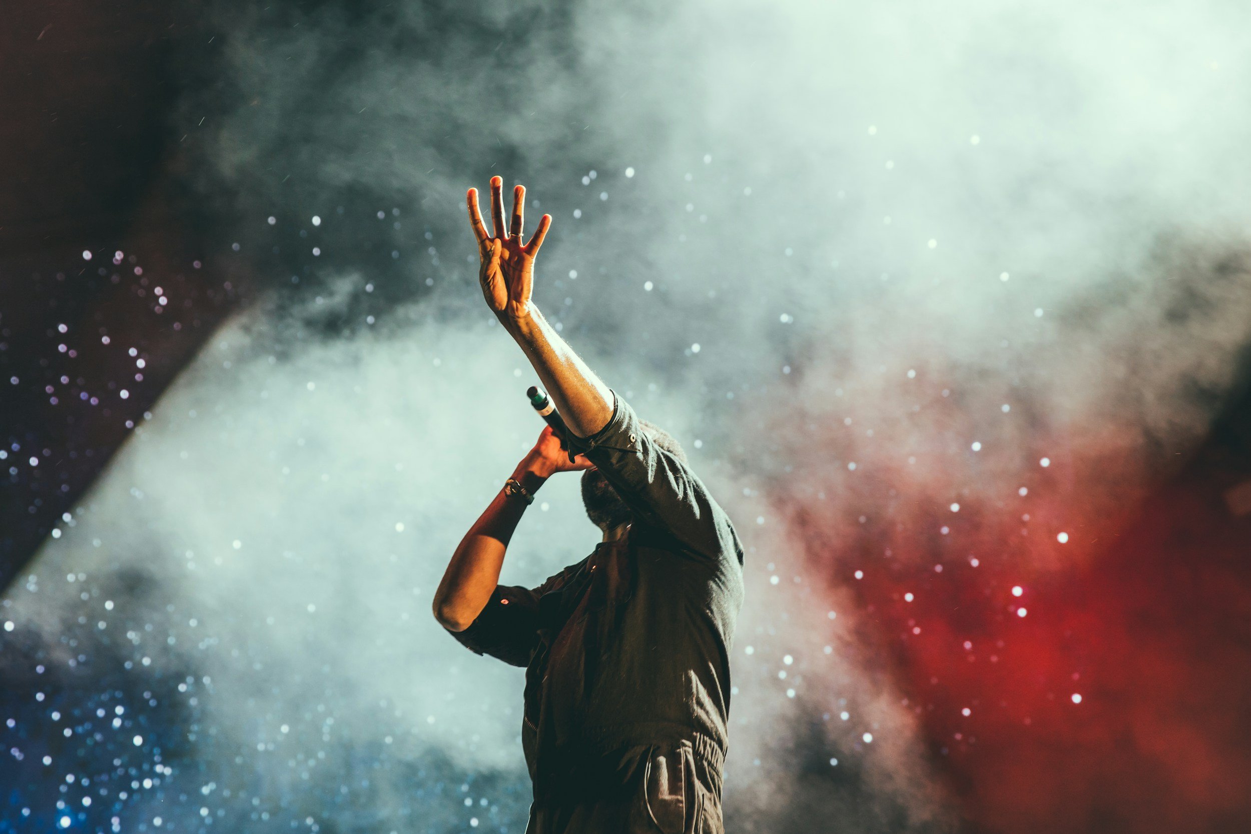 Performer holding a microphone on stage with a colorful, smoky background, raising one hand.