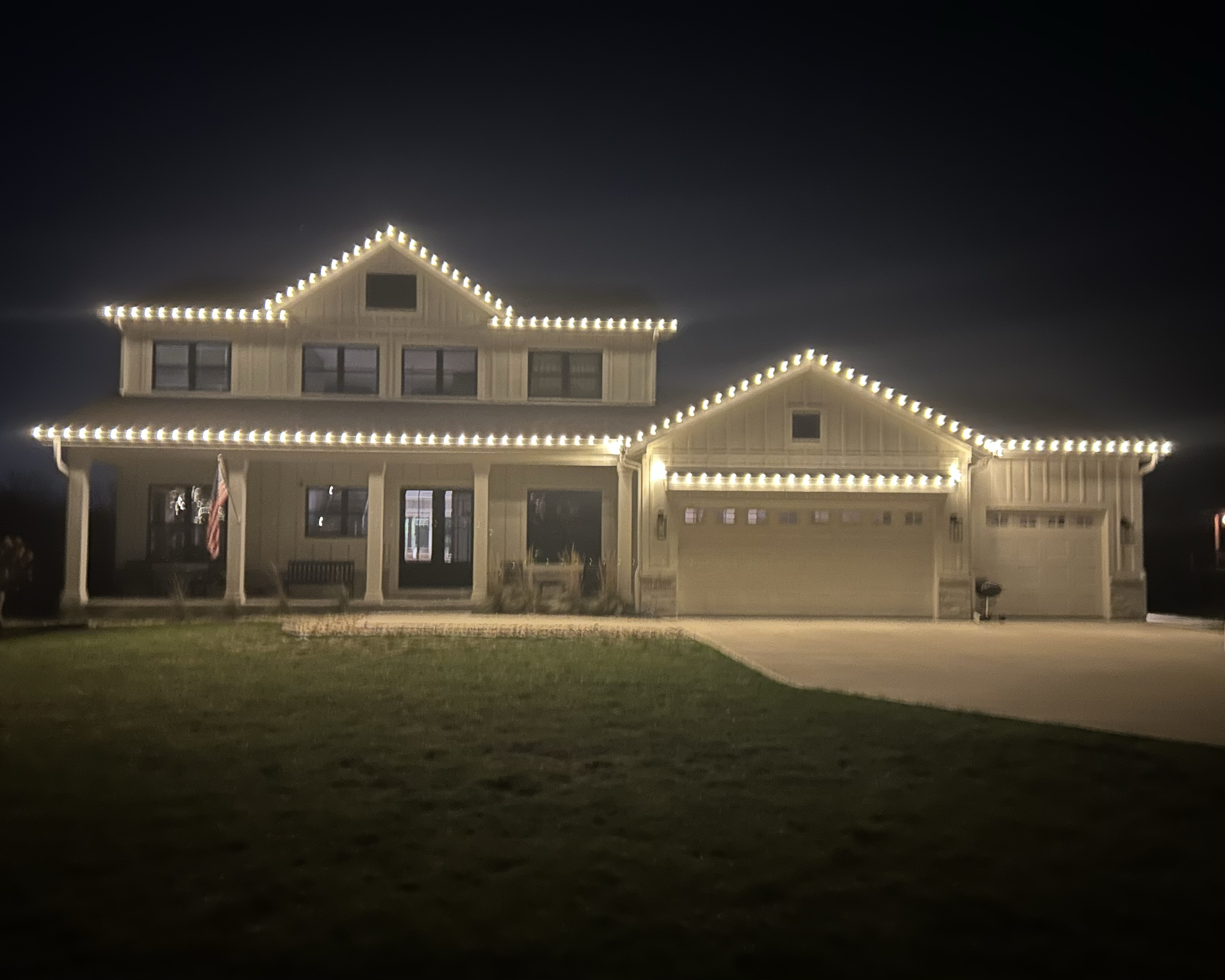 A two-story house decorated with white string lights outlining the roof and windows at night. The house has a front porch with a bench and an American flag, and a garage on the right side. There is a lawn in the front yard and a driveway leading to t