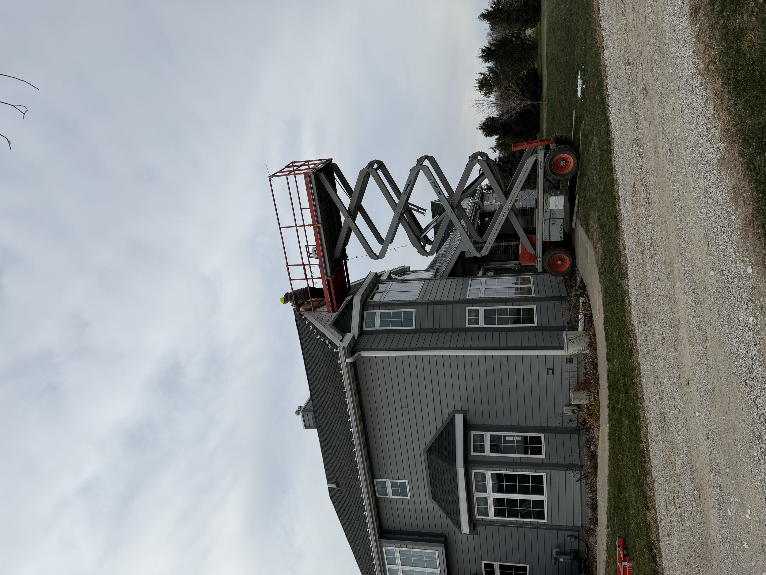 A worker in a yellow safety helmet on a red scissor lift working on the roof of a gray house.