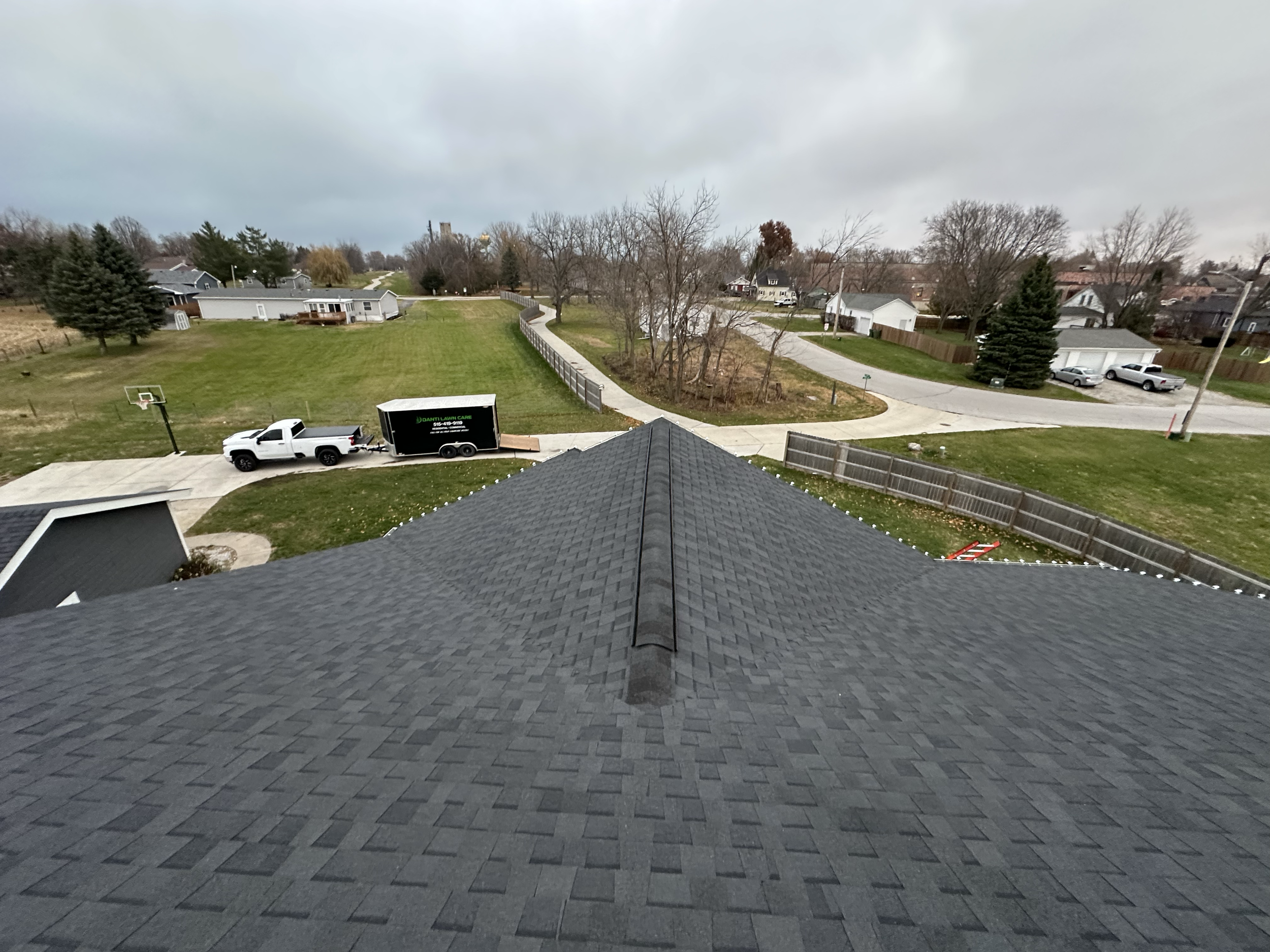 A view from a rooftop looking over a suburban neighborhood with a green backyard, a parked truck with a trailer, surrounding houses, and a cloudy sky.