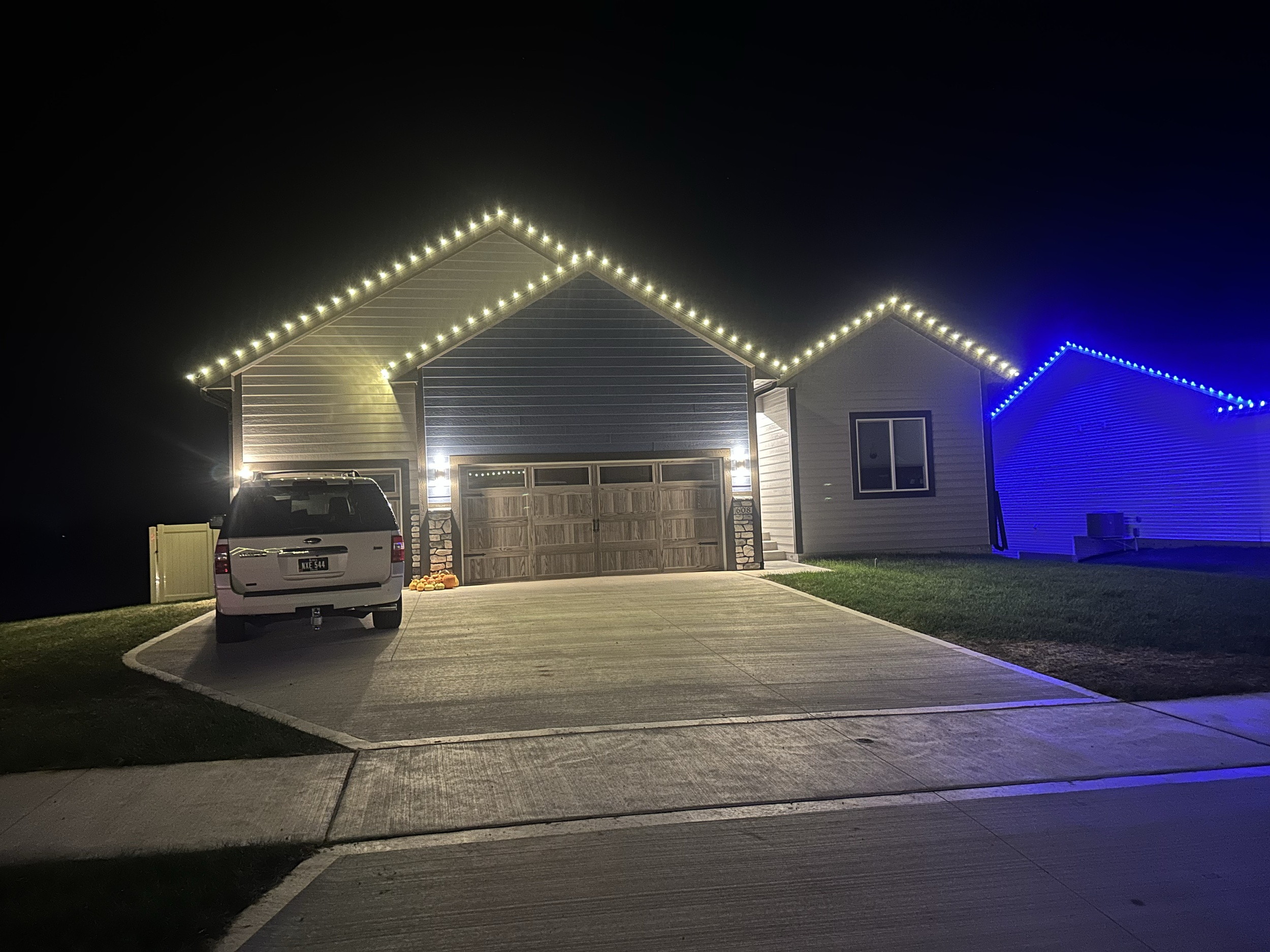 Nighttime view of a house with Christmas lights outlining the roof, a white vehicle parked in the driveway, and a neighboring house with blue lights.