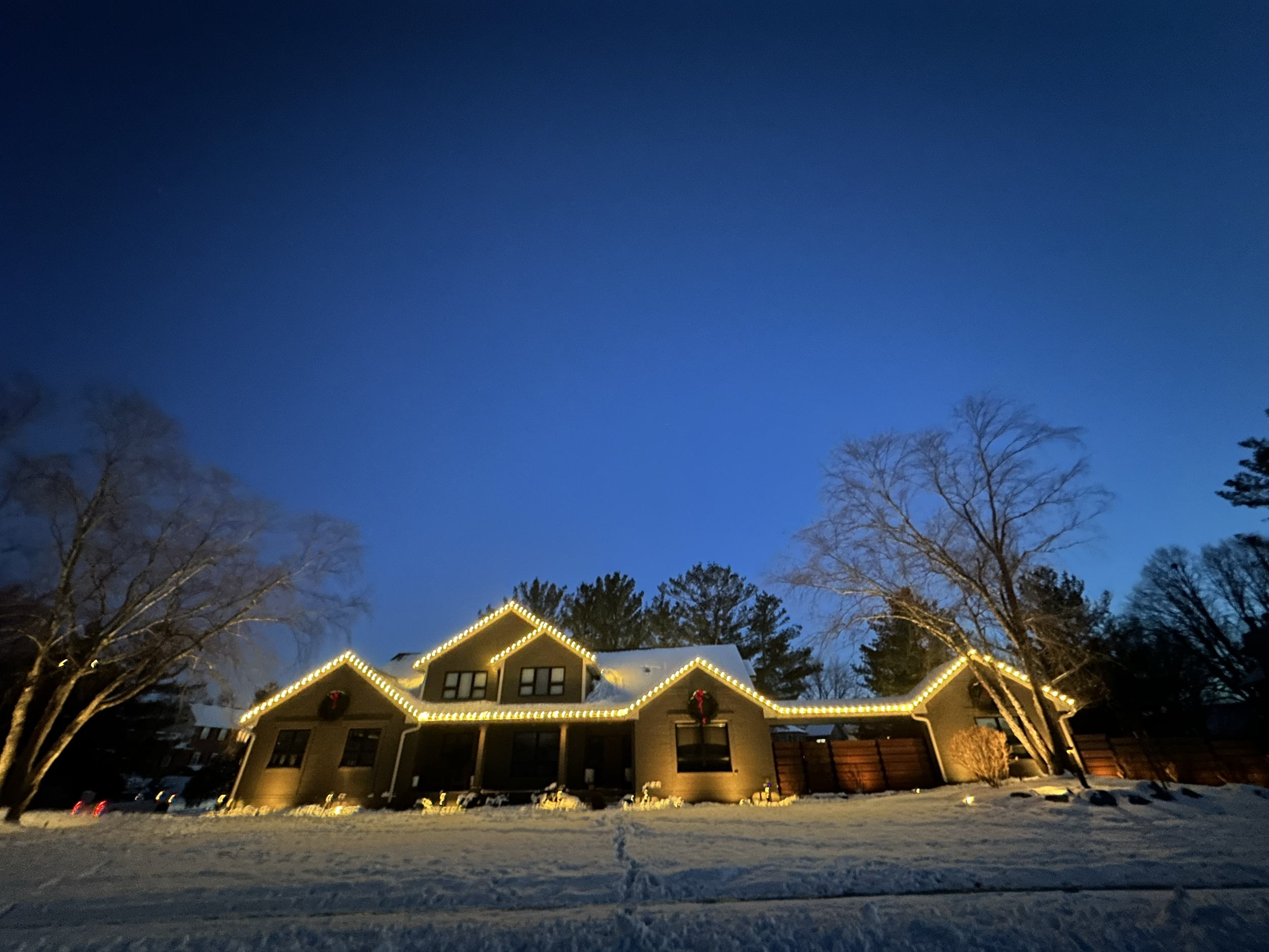 House decorated with Christmas lights and wreaths in a snowy neighborhood at dusk.