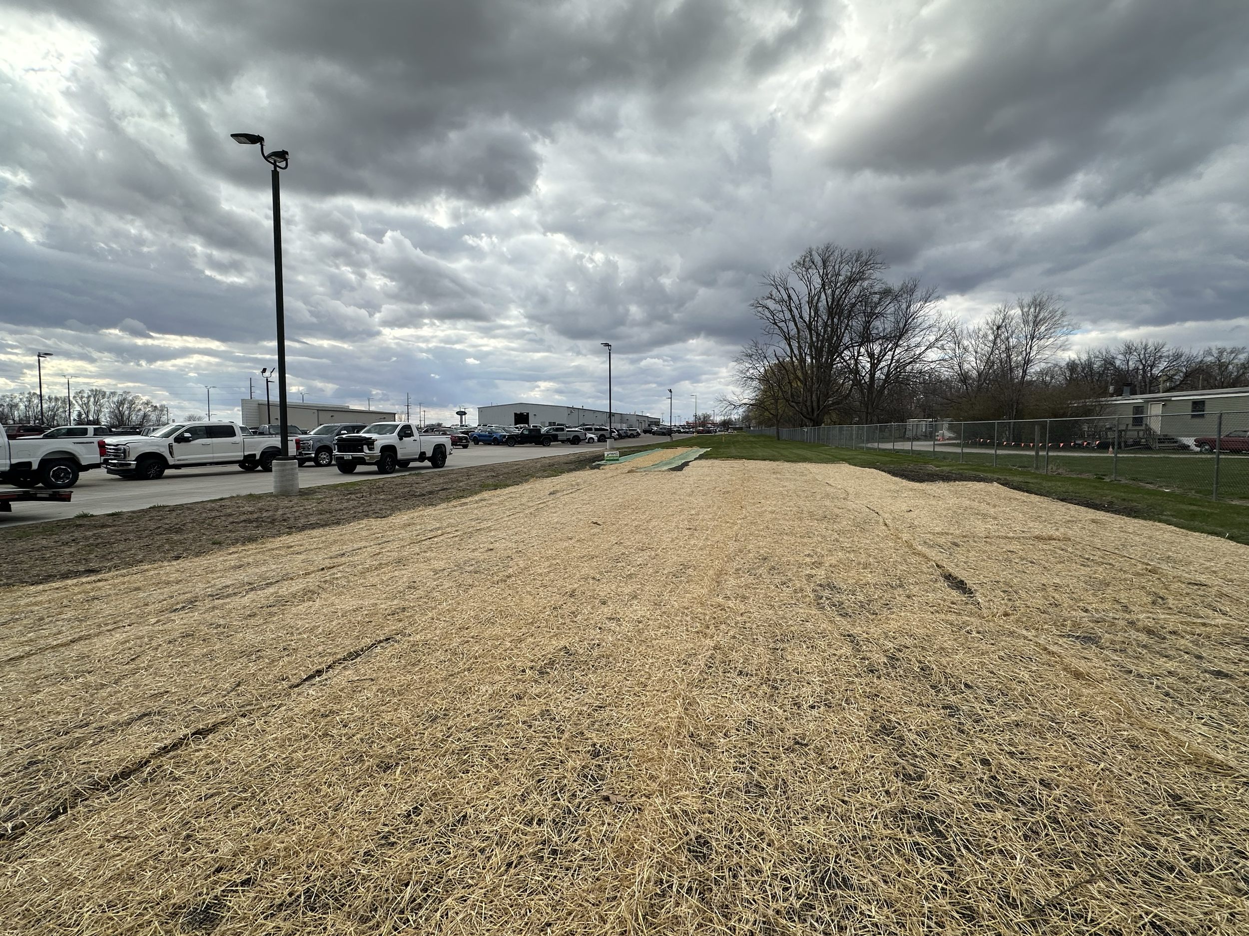 A small, leveled area covered with straw, adjacent to a parking lot with multiple white trucks parked in a row. There are tall streetlights and a chain-link fence on the right side, with leafless trees and industrial buildings in the background under
