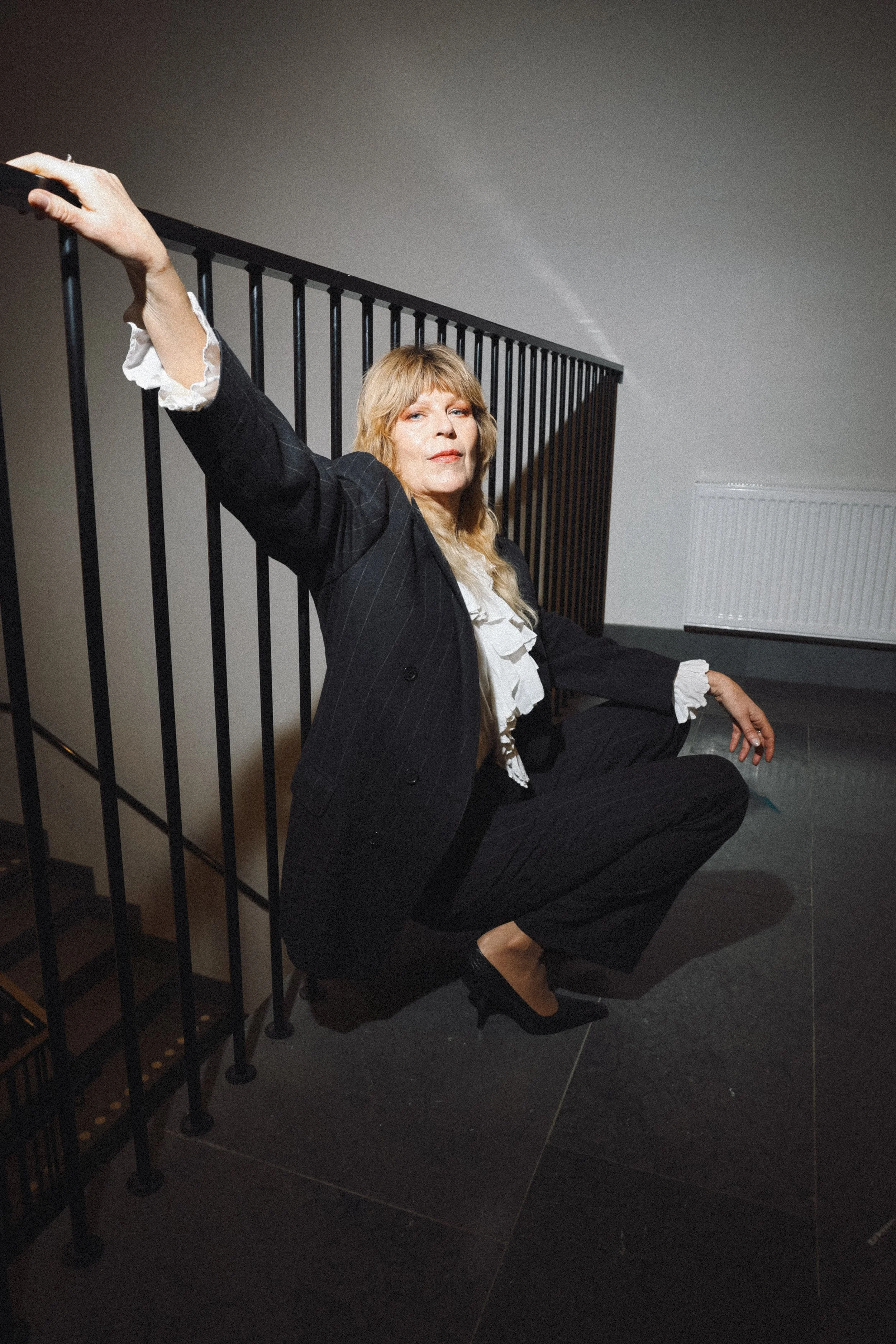 A woman in a black suit with white ruffles at the cuffs and neck, wearing high heels, crouches next to a black metal staircase railing in a dimly lit room with neutral walls and a radiator in the background.