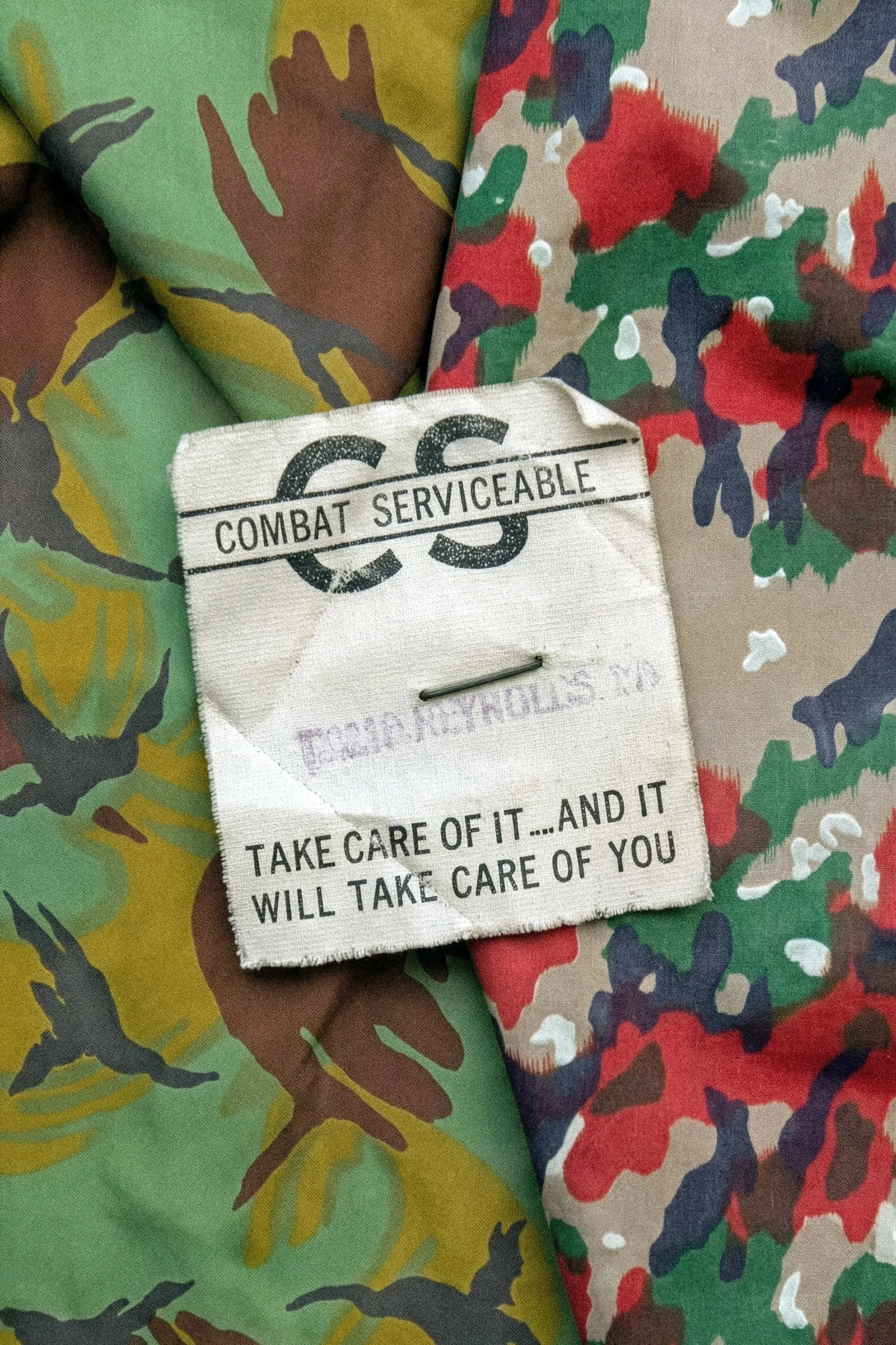 A fabric patch with the words "C.C.S. COMBAT SERVICEABLE" and a cautionary message, placed on camouflage patterned fabric.