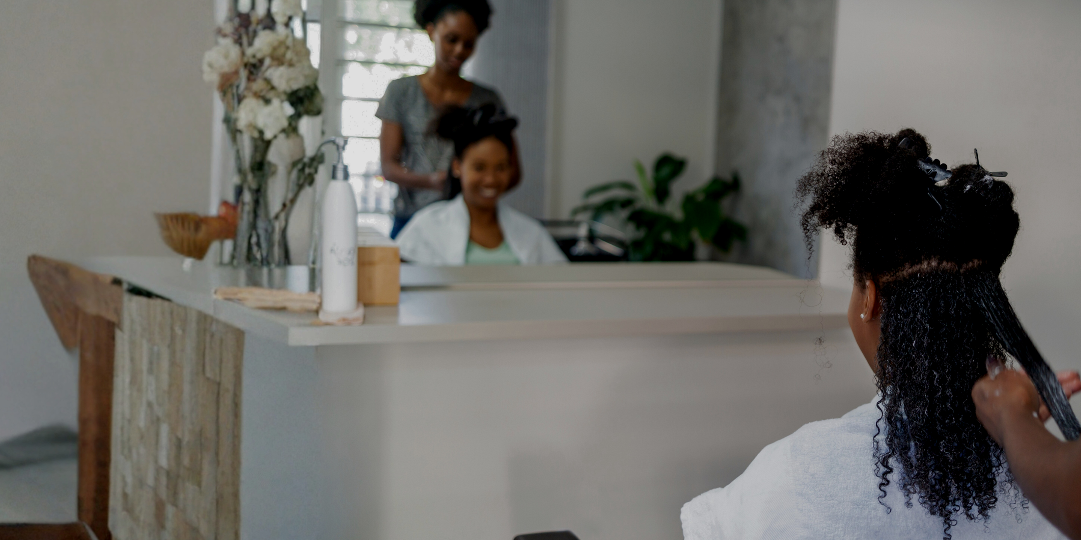 A woman getting her hair styled by a hairstylist, with another woman smiling in the background in a bright salon with plants and flowers, and a purple text box overlaying the image.