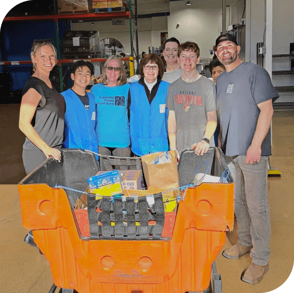 Group of seven people standing behind a shopping cart filled with food donations inside a warehouse.