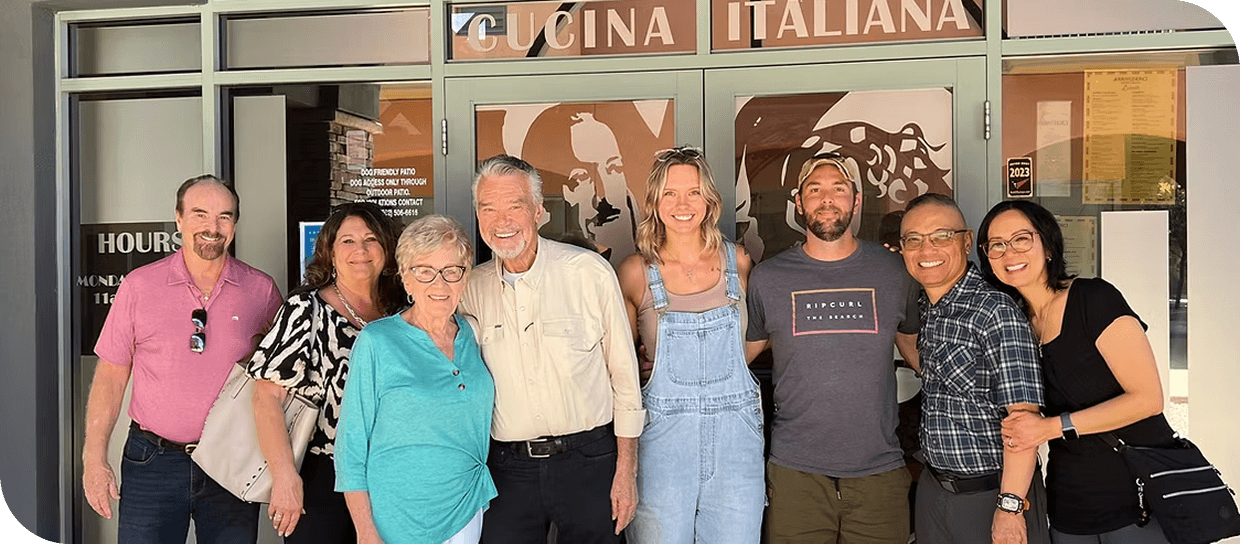 Group of nine people standing outdoors. They are smiling and posing for the photo, with some wearing casual clothing and sunglasses.