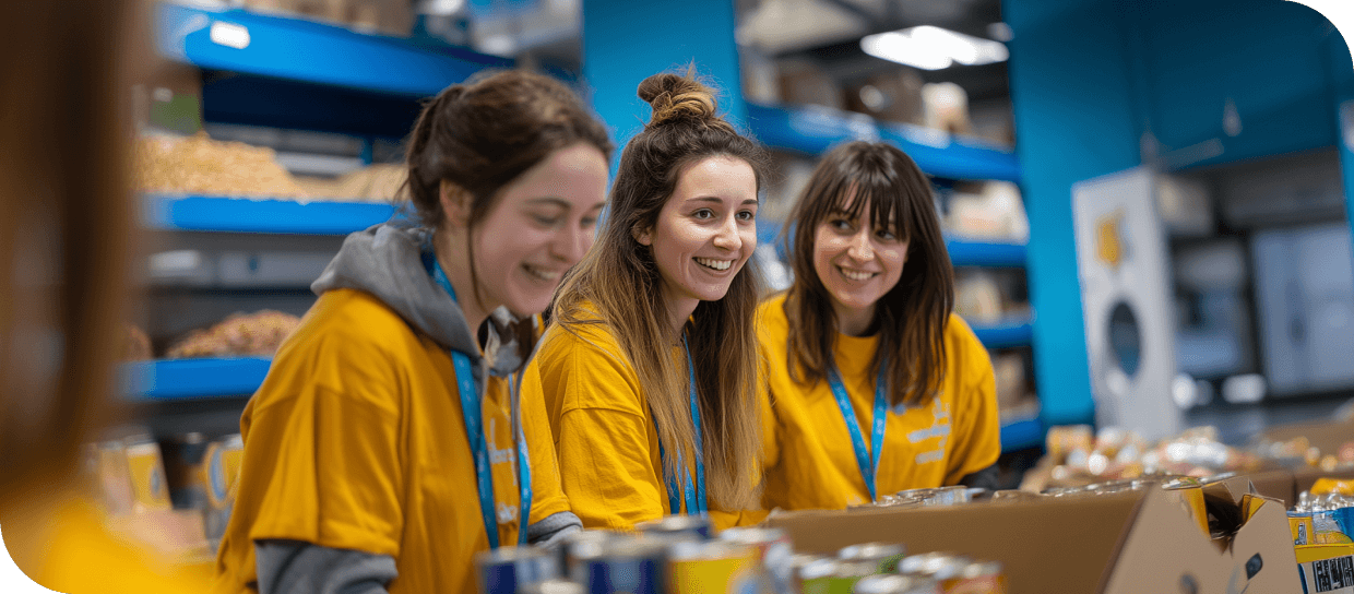 Three young women with yellow shirts and blue lanyards smiling at a food bank counter with goods.