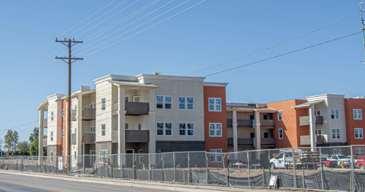 Multi-story apartment building under construction with a fenced sidewalk and parked cars, power lines visible against a clear blue sky.