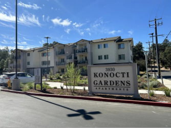 Apartment complex named Konocti Gardens with a sign displaying the address 3930, surrounded by landscaping, under a blue sky with some clouds.