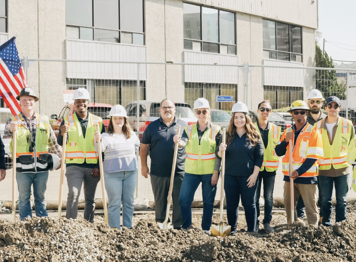 Group of construction workers and officials in safety vests and helmets at a groundbreaking ceremony, with shovels in hand, in front of a building and construction site.