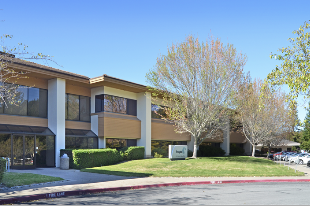 Exterior view of a two-story office building with trees and parked cars in front and a clear blue sky.