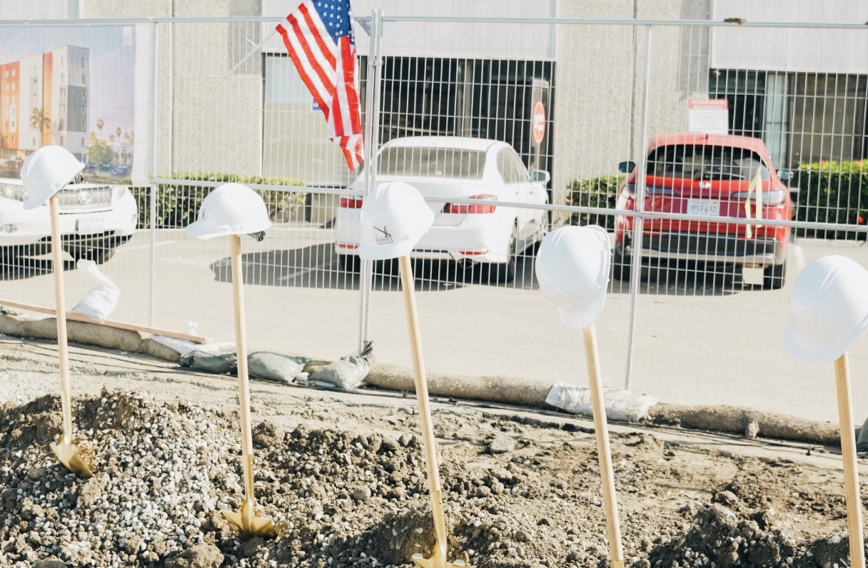 Construction site with five white safety helmets on sticks, gravel on ground, and American flag, with parked cars and a fence in the background.