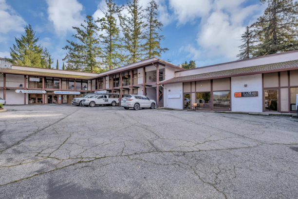 A two-story motel with a parking lot filled with cars, surrounding tall pine trees, and a partly cloudy sky.