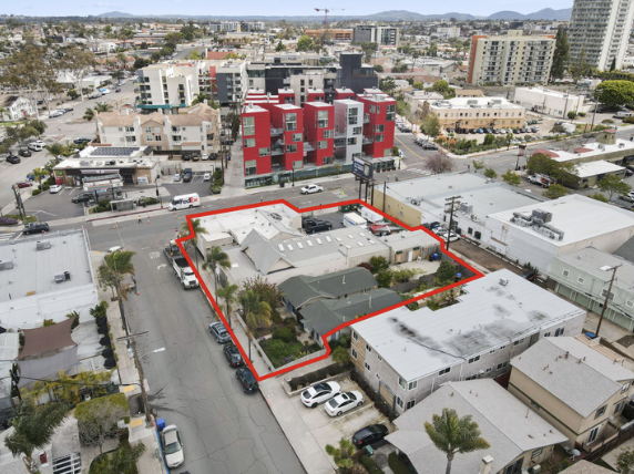 Aerial view of a city block with a red outline around a property including a house and trees, surrounded by various commercial and residential buildings, with streets and parked cars.