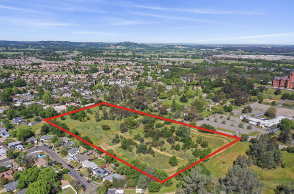 Aerial view of an open green lot with trees, outlined in red, in a suburban area with residential houses and cityscape in the distance under a blue sky.