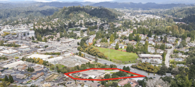 Aerial view of a suburban neighborhood with a red outlined property in the foreground, surrounded by houses, roads, and green spaces with hills in the background.