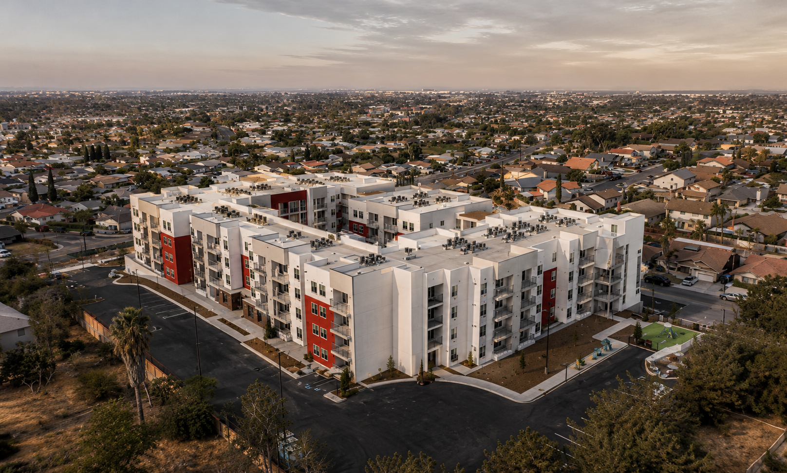 An aerial view of a large modern apartment complex with white and red exterior, surrounded by parking lots, residential houses, and some trees under a cloudy sky at dusk.