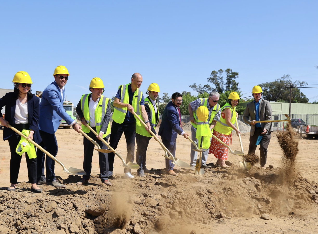Group of people in business attire and construction vests with yellow hard hats, participating in a groundbreaking ceremony on a construction site, shoveling dirt.