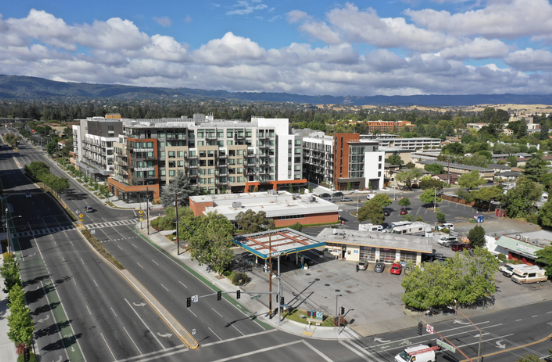 An aerial view of a mixed-use urban area showing modern residential buildings, a parking lot, trees, and surrounding streets under partly cloudy skies.
