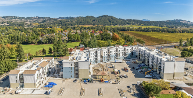 Construction site of a multi-story residential complex with surrounding parking area and scenic green landscape with hills in the background.