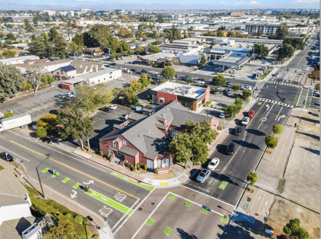 Aerial view of a city street corner with a red building, parked cars, trees, and surrounding commercial buildings.