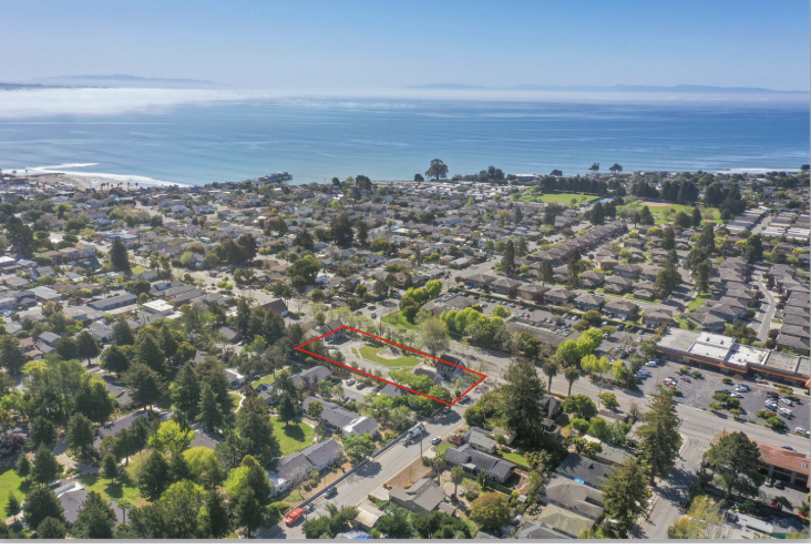 Aerial view of a coastal residential neighborhood with a vacant lot outlined in red, near the beach and ocean, surrounded by trees and houses.
