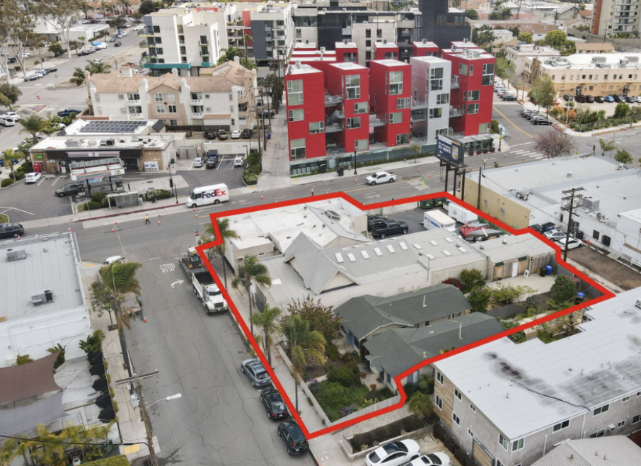 Aerial view of a city block with a residential property outlined in red, surrounded by various commercial and residential buildings, parked cars, and a street with traffic.