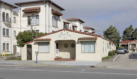 A small, single-story white building with a red tile roof, situated on a street corner with a tree and parked cars nearby.