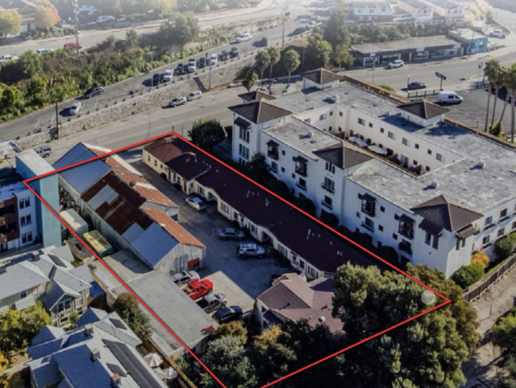 Aerial view of a property outlined in red, including a parking lot with cars, a small building, and surrounding residential buildings and trees.
