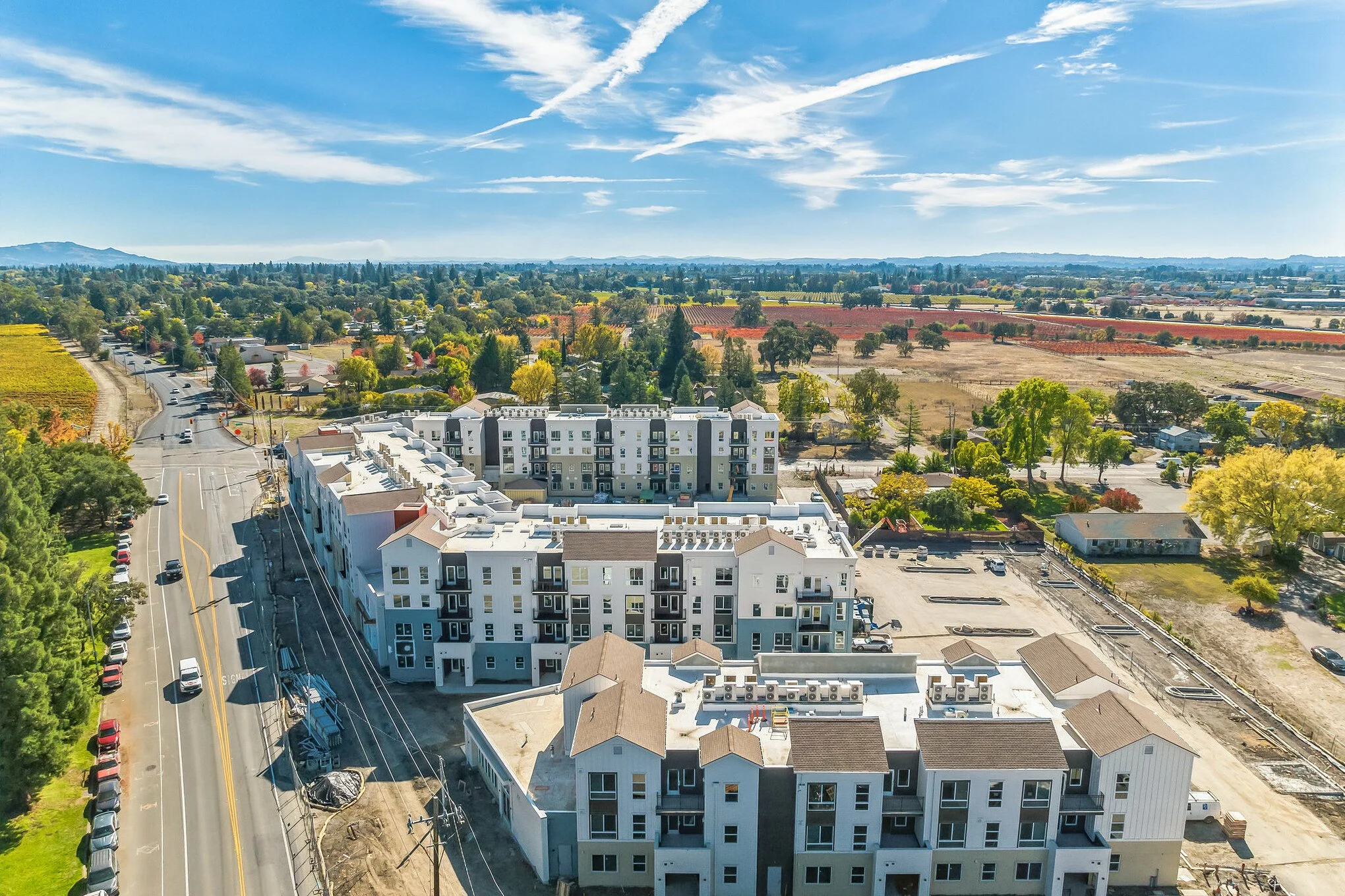 An aerial view of a cityscape with a row of white residential buildings in the foreground, a street with parked cars on the left, and lush green trees and open fields stretching to the horizon. The sky is bright blue with scattered clouds and contrails.