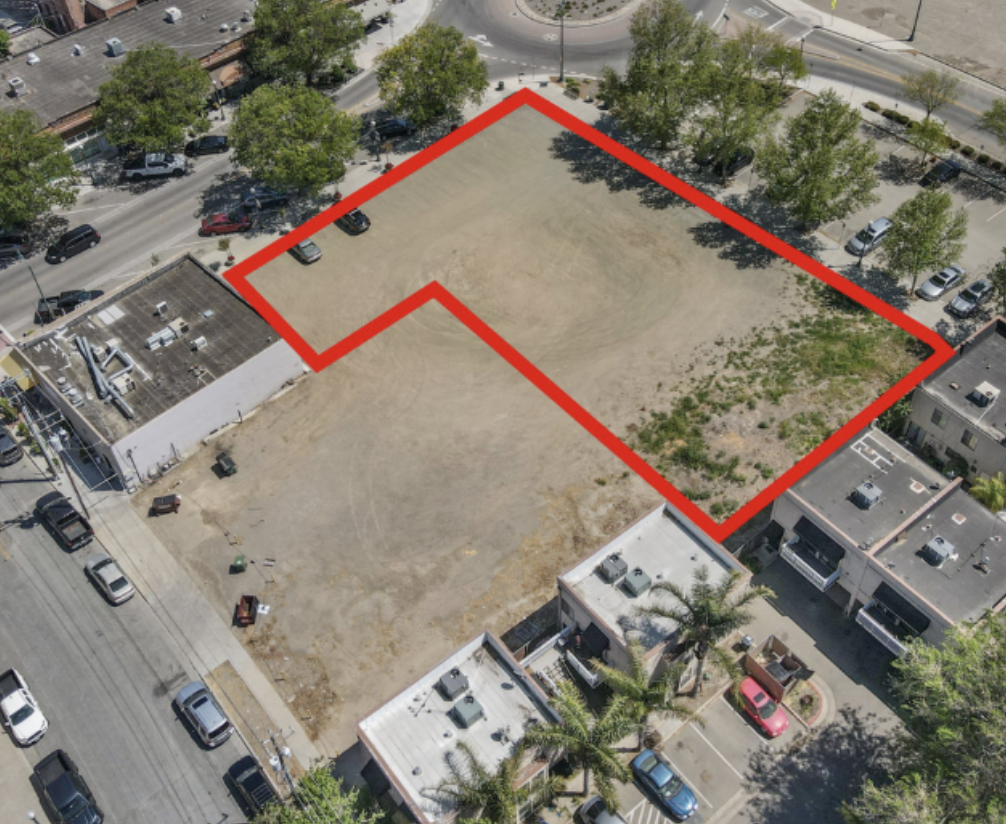 An aerial view of a vacant lot outlined in red, with surrounding buildings, parked cars, and trees.