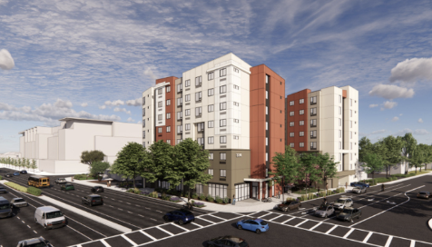 Modern mid-rise apartment building on a city street corner with a parking lot and trees, under a partly cloudy sky.