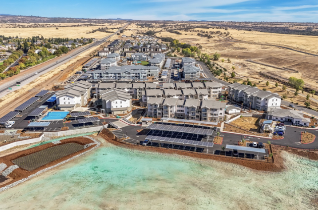 Aerial view of a newly built residential complex near a body of water, with solar panels on rooftops and a parking lot, surrounded by open land and a highway in the distance.