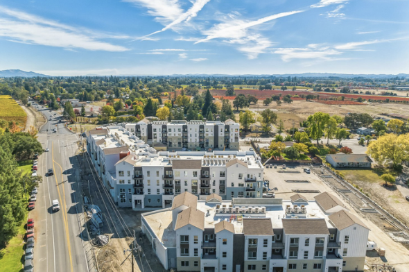 Aerial view of a modern apartment complex with multiple buildings, parking lot, and surrounding landscape under a clear blue sky.