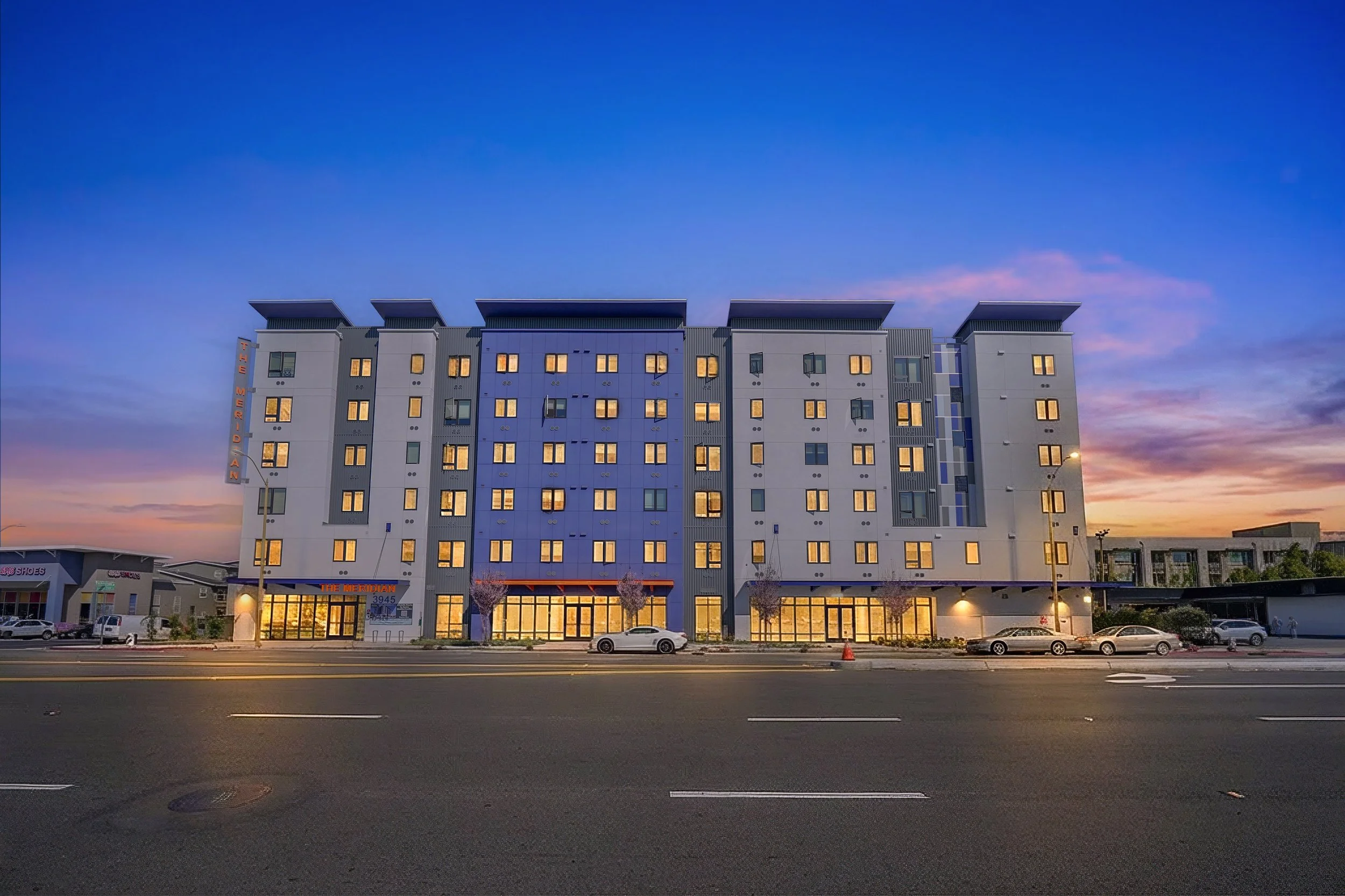 A multi-story modern hotel building named The Meridian with illuminated windows at dusk. The hotel has a sign on the left side, several vents on the roof, and a parking lot in front with a few cars and small trees. The sky is a gradient of deep blue, purple, and orange.