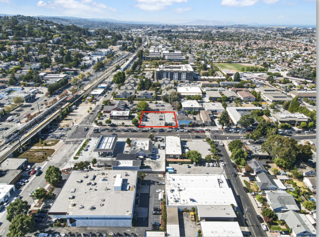 Aerial view of a city street with buildings, trees, and parking lots, showing a highlighted parcel of land outlined in red.