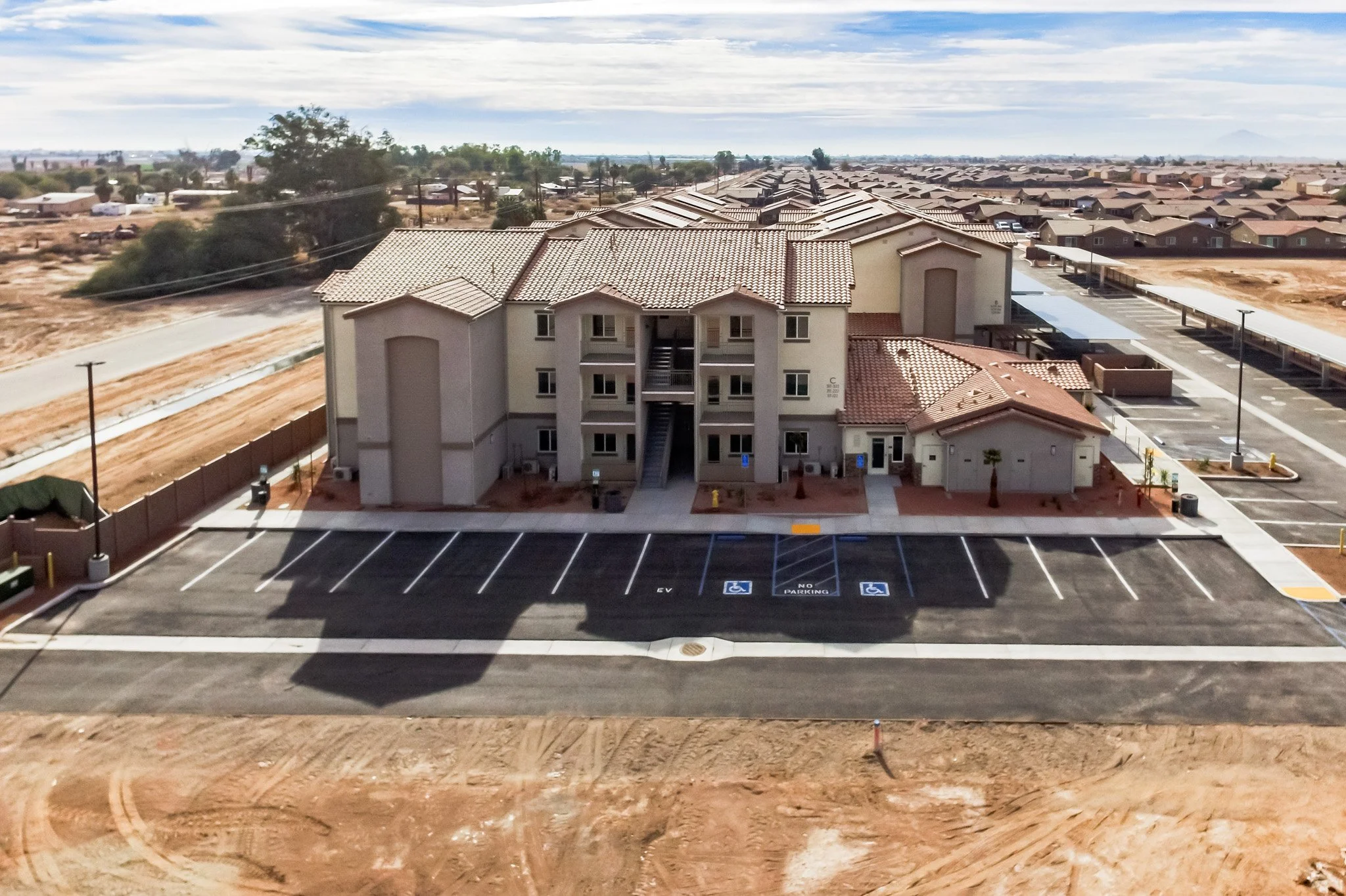 A multi-story residential building with a tiled roof and staircases outside. In front, a parking lot with marked handicapped spots and a sign for electric vehicle charging stations. Surrounding the building are bare patches of land and underdeveloped areas, with a suburban neighborhood in the background and a partly cloudy sky overhead.