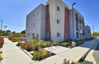 Modern apartment building with landscaped garden in front, concrete sidewalk, and blue sky.