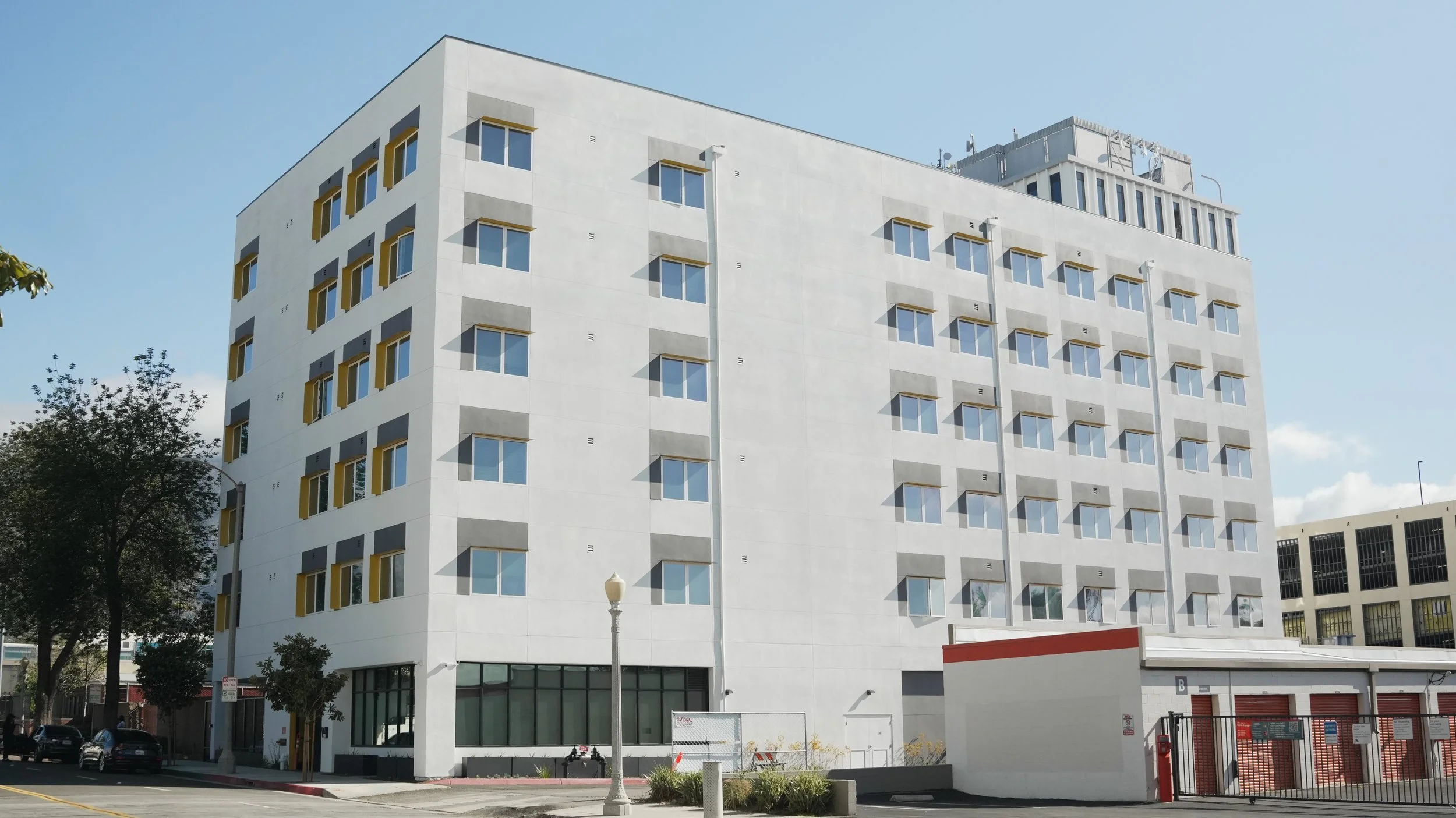 A modern white multi-story building with multiple rectangular windows, some with yellow trim, under a partly cloudy sky. Trees and street elements are in the foreground.