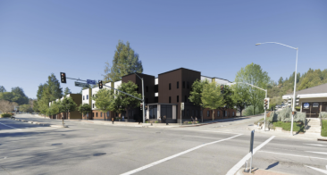 An urban intersection with traffic lights, crosswalks, and a modern building on the corner, surrounded by trees and a clear blue sky.